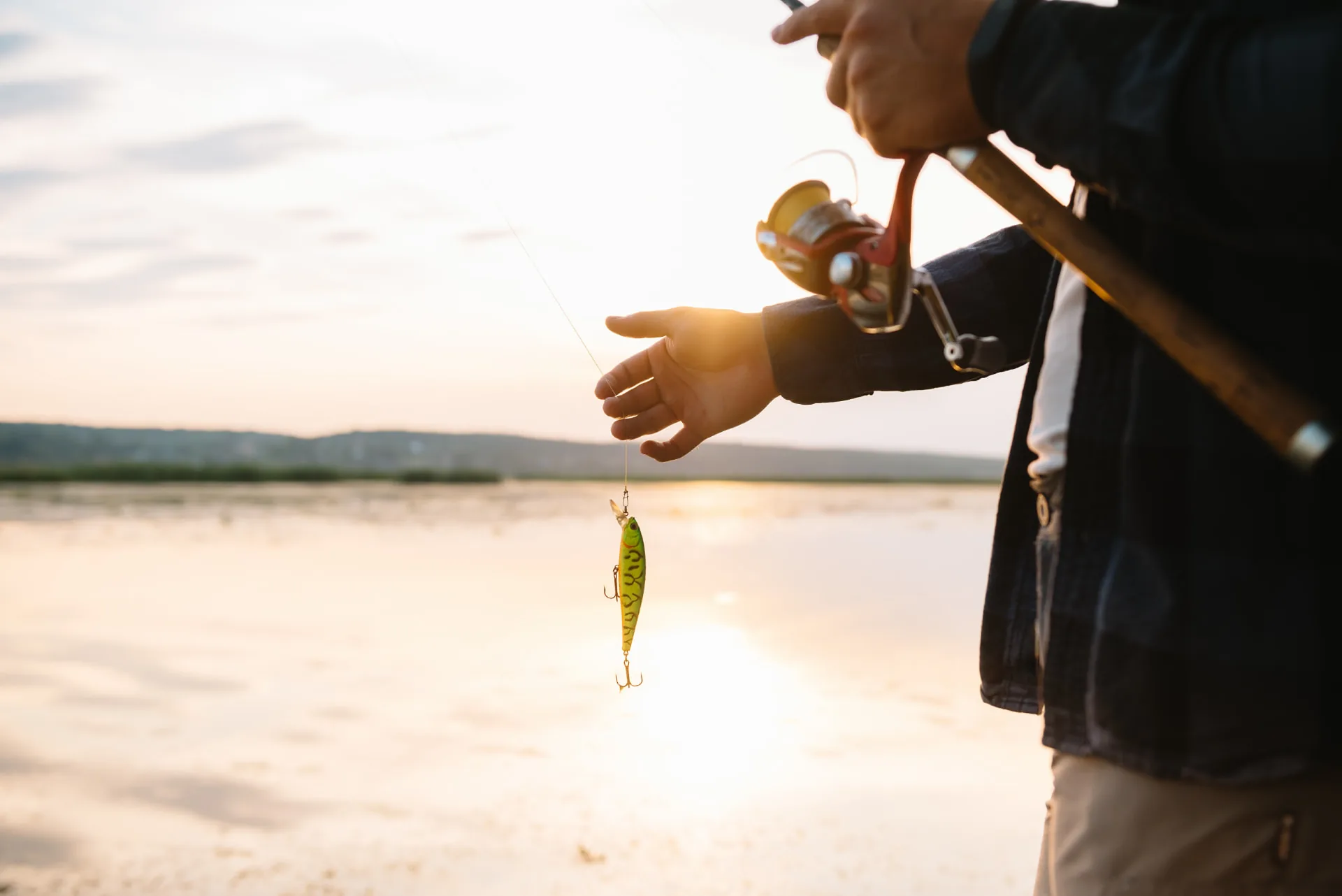 Fisherman holding a spinning rod at golden hour, a lure hanging from the line over calm water